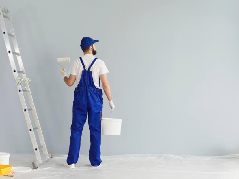 A worker standing against wall holding paint roller and paint on floor covered with Plastic sheet In Essex UK